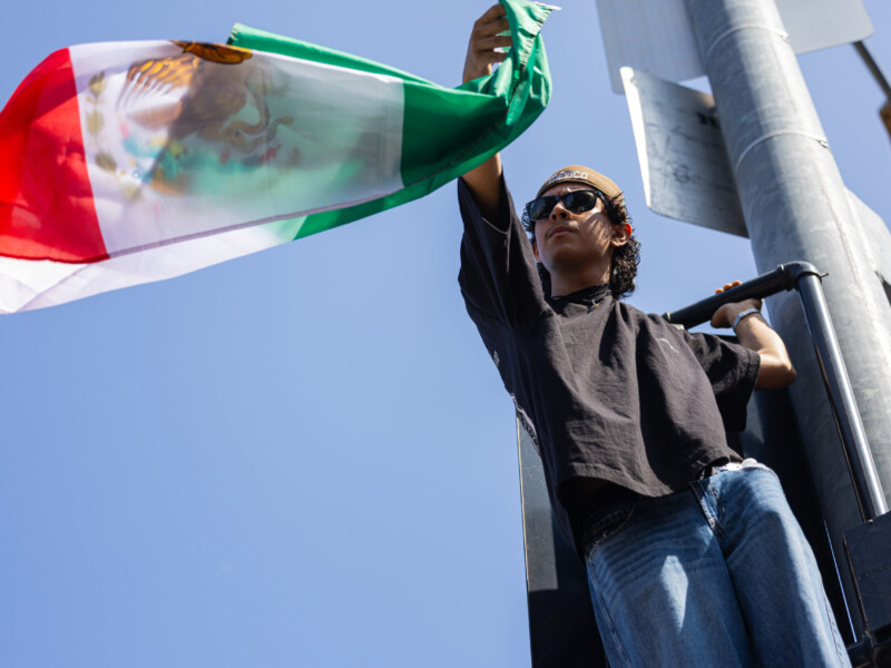 An anti-ICE protester waves a Mexican flag while hanging on a street light post at Temple and Alameda Streets in downtown Los Angeles on June 8. Police broke up the protests as demonstrators clashed with officers. Ethan Cohen | Long Beach Current